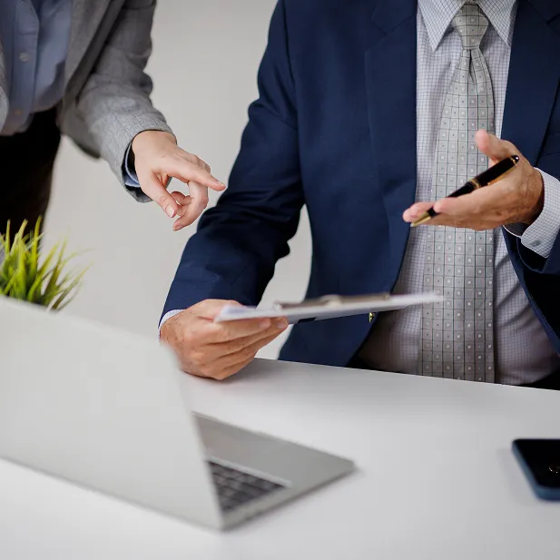 Two colleagues review documents on a clipboard together — one in a blue suit gestures with a pen while another in a gray blazer points at the papers.