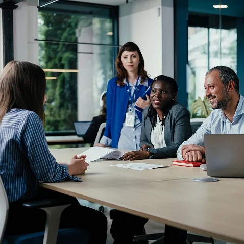 Four diverse professionals gathered around a conference table with a laptop and red notebook, smiling in a bright office with greenery outside.