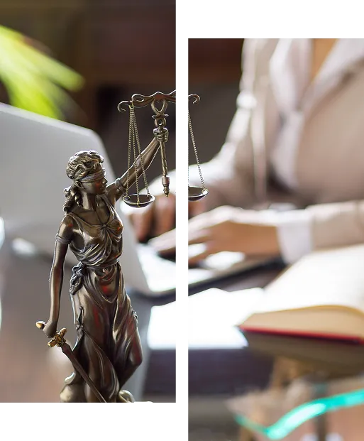 A bronze Lady Justice statue with balanced scales and a sword, blurred in the background is a person typing at a laptop beside an open book.