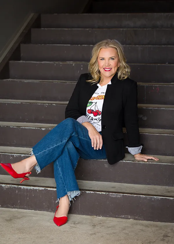 Molly in a black blazer, cherry-print white tee, rolled-up jeans, and red heels, sitting on dark gray steps with her ankles crossed.