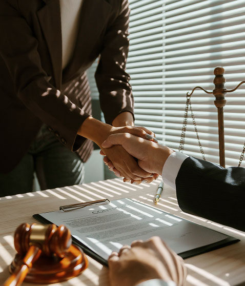 Two individuals in suits shake hands across a table where a document, pen, and pair of glasses lie, with a wooden gavel and a scale of justice nearby. Sunlight streaming through blinds casts horizontal shadows across the scene.